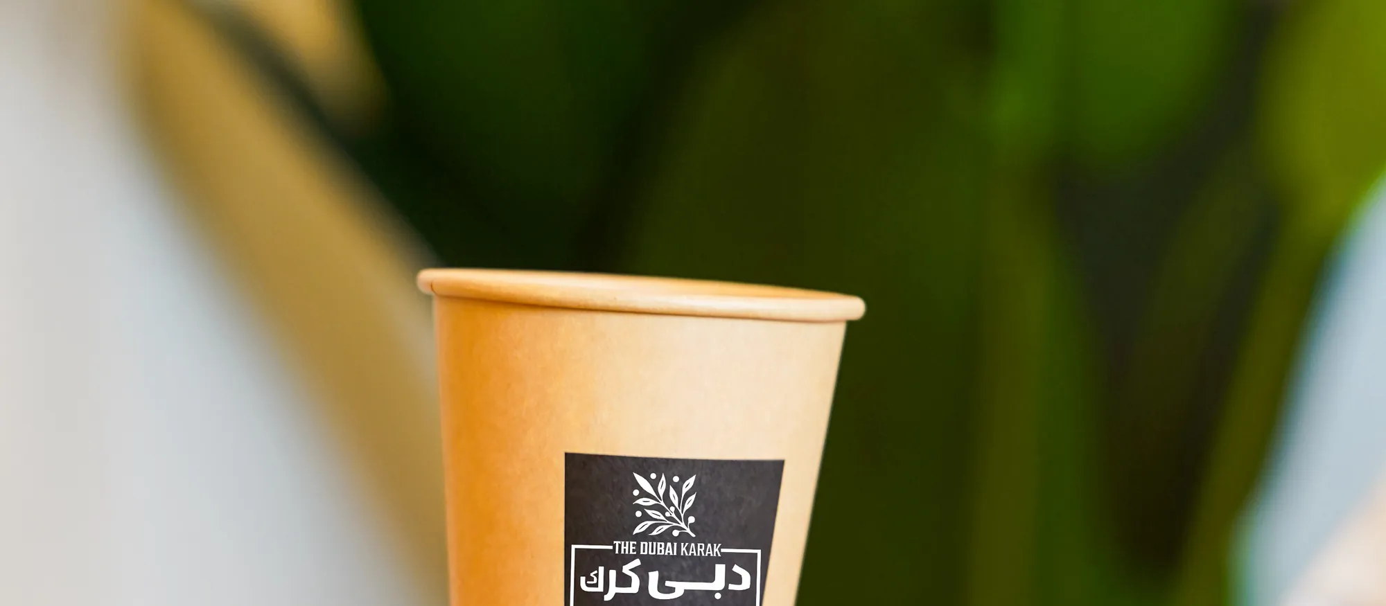 A branded cup of Dubai karak chai held in the palm of a hand beside tropical greenery